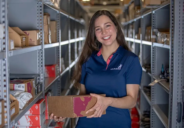 Asesora sonriendo mientras sostiene una caja de repuestos en el almacén de refacciones Mack Trucks.